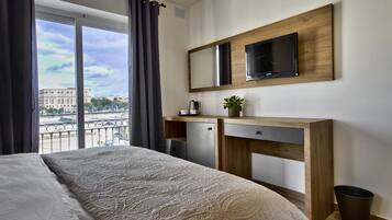 A modern hotel room with a large bed, a wooden desk, a flat-screen TV, and a view of a cityscape through large windows.