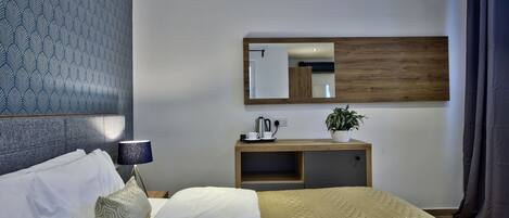 Quarto de hotel com cama, almofadas, mesa de cabeceira com abajur, espelho, cabeceira de madeira, planta e janela com cortinas.