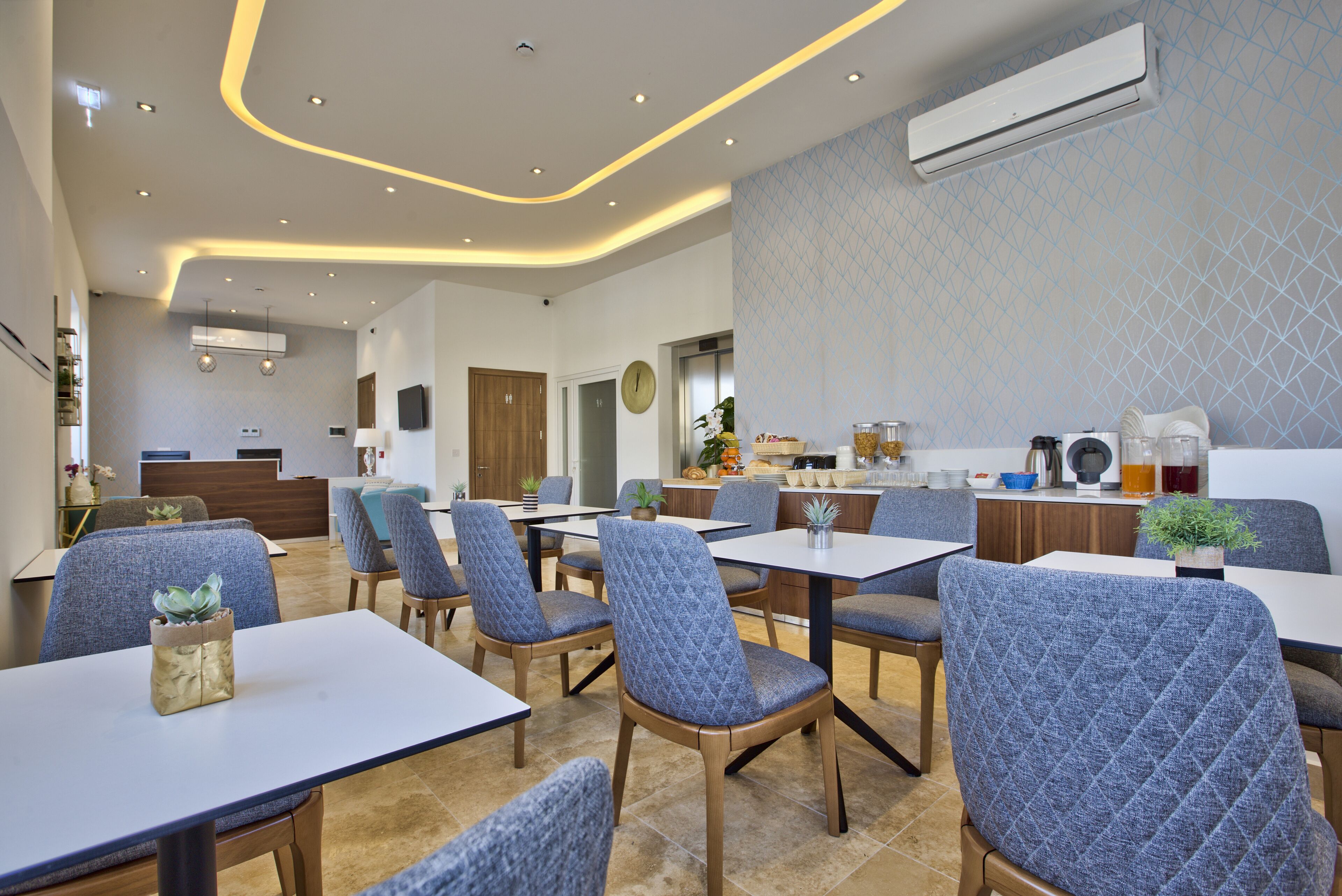 A modern restaurant interior with blue upholstered chairs, white tables, and a kitchen area in the background.
