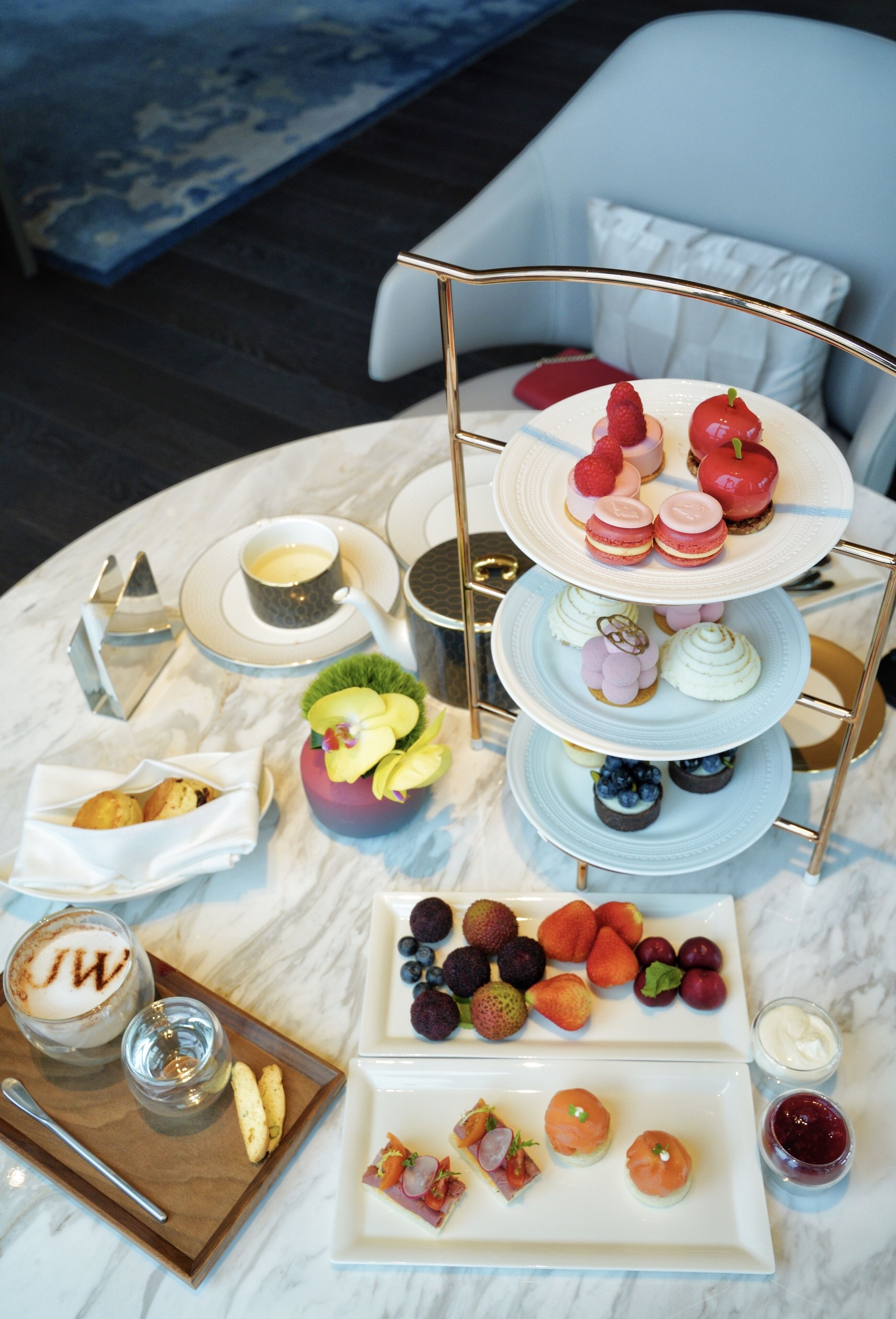 A multi-tiered dessert stand with various pastries and fruits on a marble table, accompanied by small plates of snacks and a tray with a drink and a dessert.