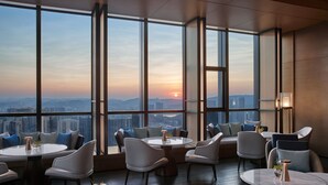 Un interior moderno con mesas y sillas redondas, grandes ventanales que ofrecen vistas a la ciudad, y un atardecer visible a través del vidrio.