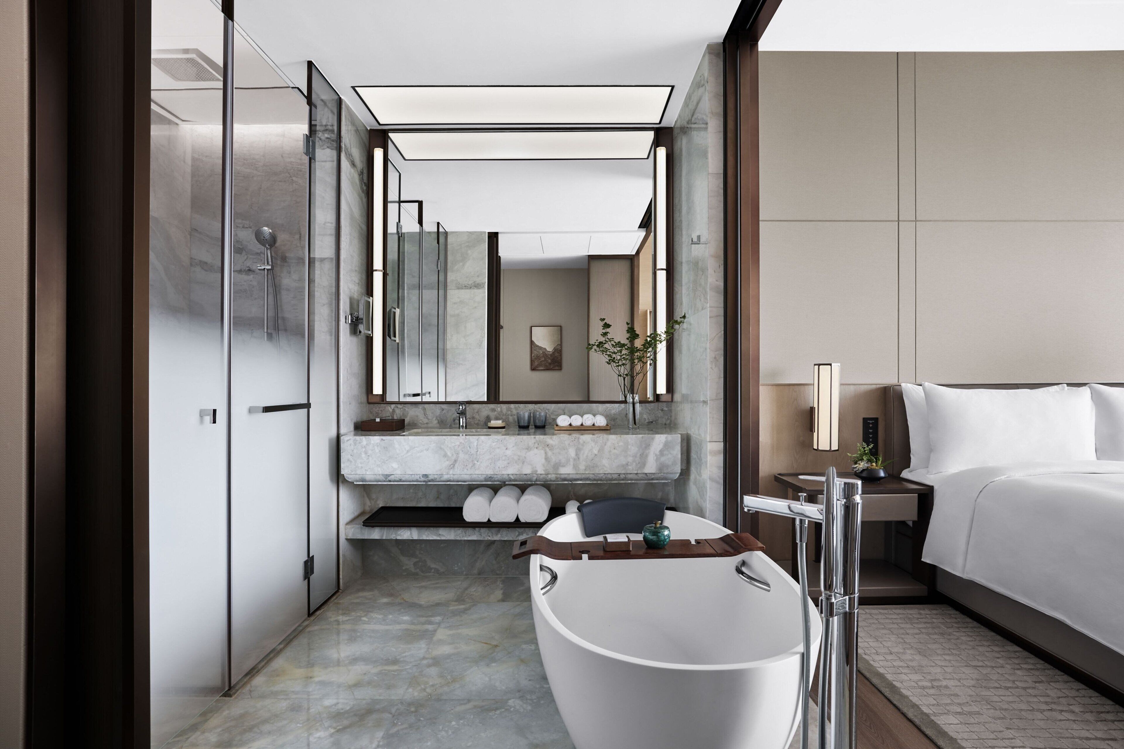 A modern hotel bathroom with a freestanding bathtub, a large mirror, a vanity with a marble countertop, and a bed with white linens.
