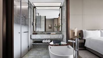 A modern hotel bathroom with a freestanding bathtub, a large mirror, a vanity with a marble countertop, and a bed with white linens.