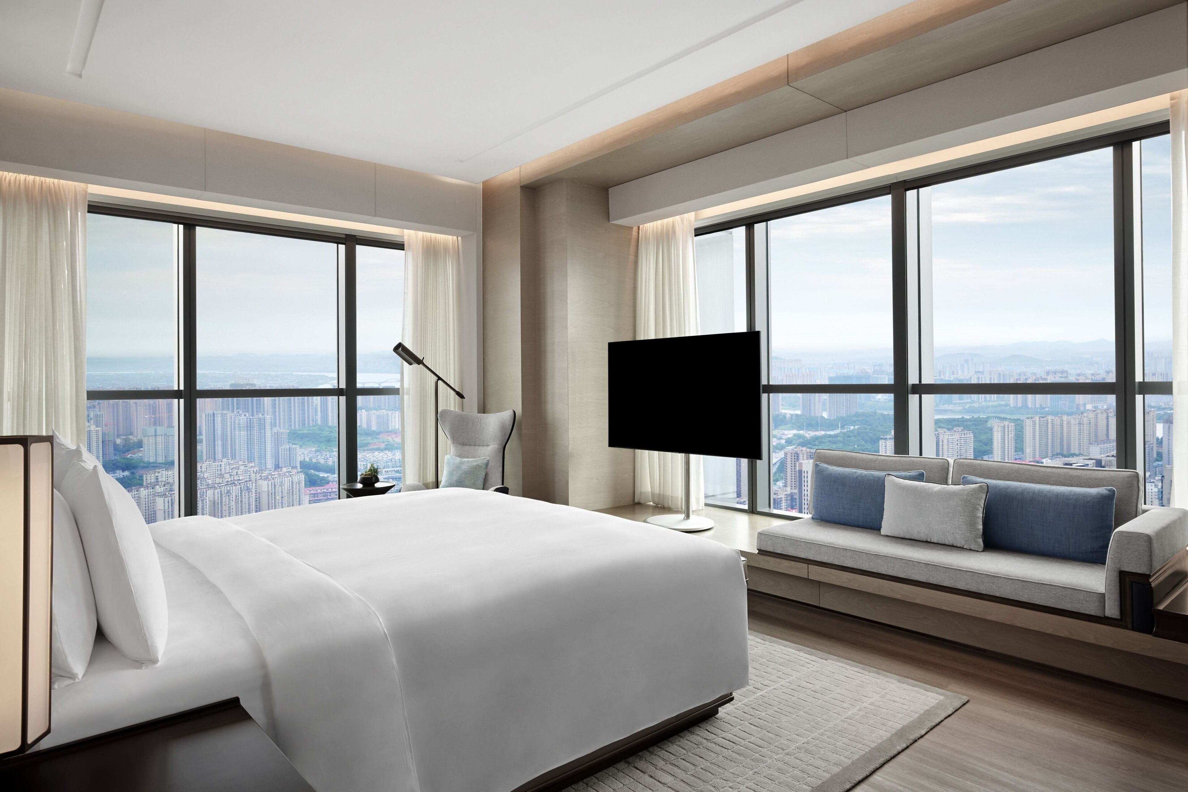 A modern hotel room with a large bed, a TV, and a sofa by a window with a city view.
