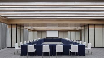 Ein Konferenzraum mit langem Tisch, weißen Stühlen und einer Projektionsleinwand.