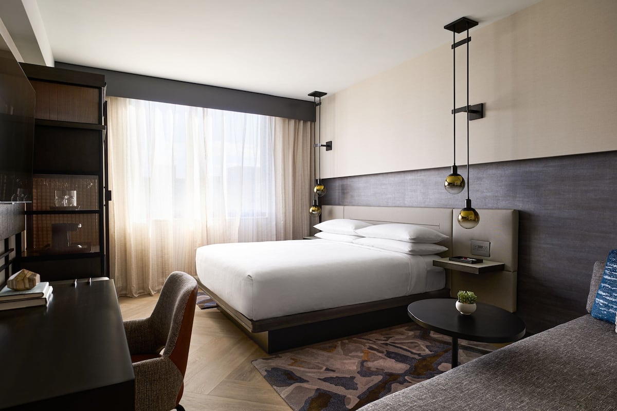 A modern hotel room with a large bed, a desk, a chair, and a small table with a plant.