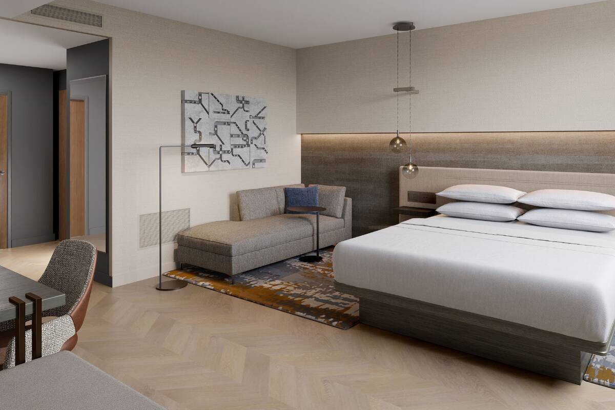 A modern hotel room with a large bed, a sofa, a desk, and a wall-mounted artwork.
