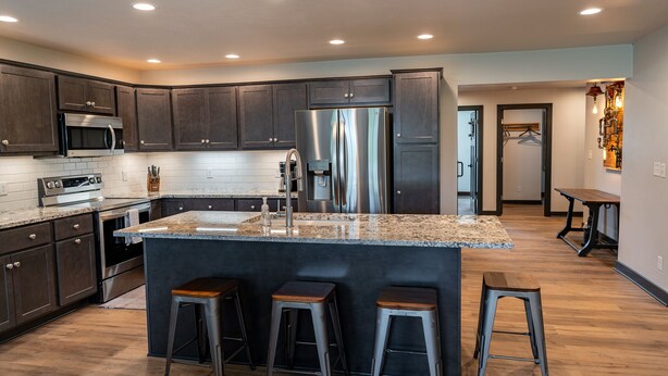 A modern kitchen with dark cabinets, stainless steel appliances, a granite countertop, and three stools. - 𝗨𝗻𝗶𝗾𝘂𝗲 𝗦𝘁𝗮𝘆 at the Carriage Haus (Manitowoc)