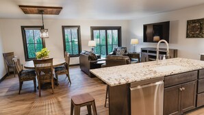 A modern kitchen with a large island, wooden dining table, and chairs. There are three windows providing views of greenery. - 𝗨𝗻𝗶𝗾𝘂𝗲 𝗦𝘁𝗮𝘆 at the Carriage Haus (Manitowoc)