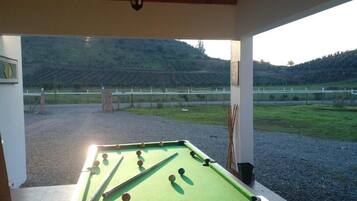 A pool table in a covered outdoor area with a view of a vineyard and hills in the background.