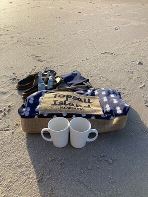 Cena de praia com uma bolsa de palha com a etiqueta 'Top of the Island N. Carolina' contendo sandálias, uma toalha e duas canecas brancas.