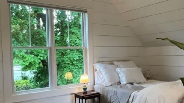 A cozy bedroom with a bed, a bedside table with a lamp, and a window with a view of greenery.