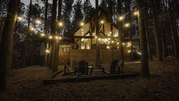 A cabin with a deck, surrounded by trees, and string lights. - ✨Live in the Moment🔥Fireplaces 💦Hot Tub 🎮Games 🛏️Luxury Suites 🌲Outdoor Fun (Broken Bow)