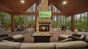 A covered deck with a stone fireplace, outdoor seating, and a television screen displaying a football game. - ✨Live in the Moment🔥Fireplaces 💦Hot Tub 🎮Games 🛏️Luxury Suites 🌲Outdoor Fun (Broken Bow)
