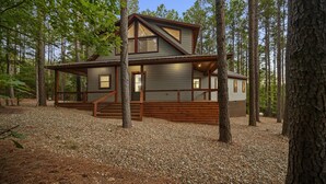 A cabin with a wooden deck, a staircase leading to the entrance, and a forested backdrop. - ✨Live in the Moment🔥Fireplaces 💦Hot Tub 🎮Games 🛏️Luxury Suites 🌲Outdoor Fun (Broken Bow)