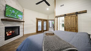 A bedroom with a fireplace, a television mounted on the wall, and a sliding barn-style door. - ✨Live in the Moment🔥Fireplaces 💦Hot Tub 🎮Games 🛏️Luxury Suites 🌲Outdoor Fun (Broken Bow)