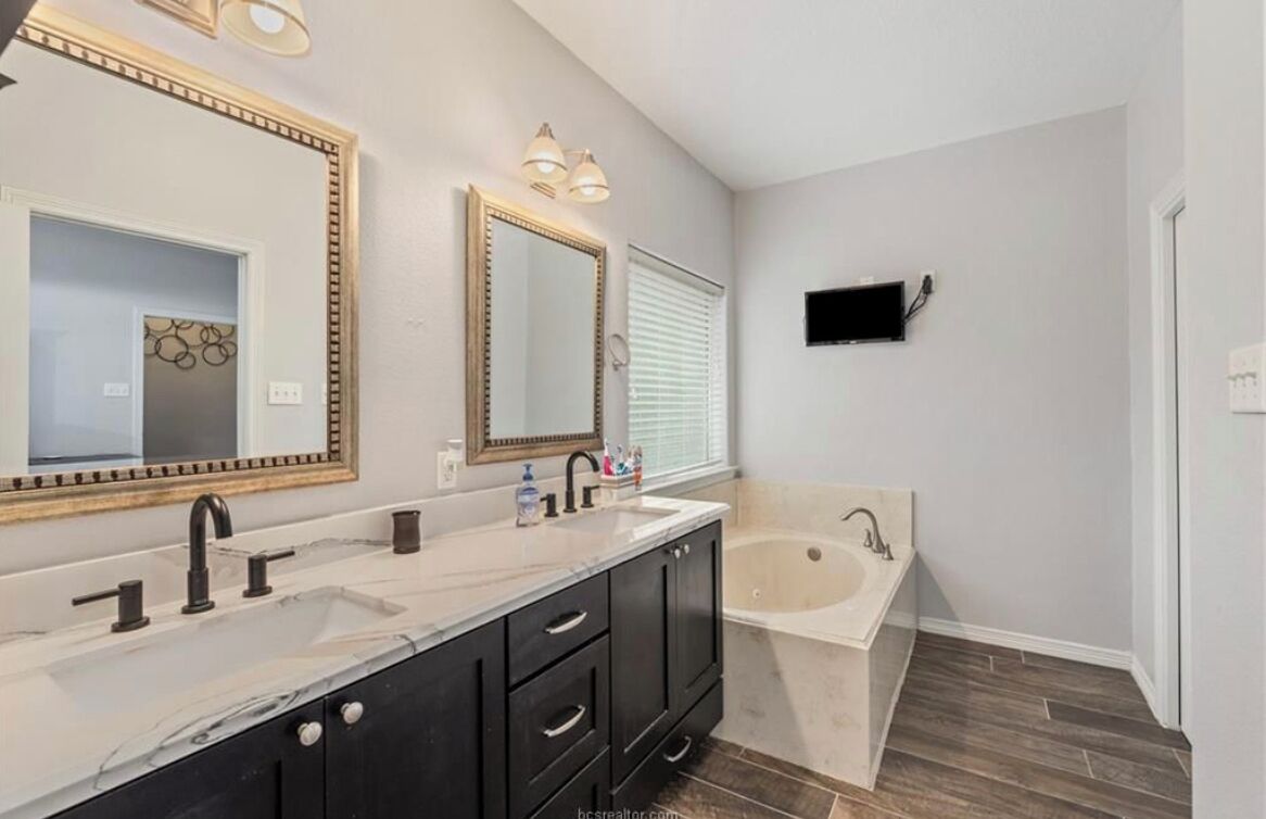 A modern bathroom with a double vanity, a large mirror, a bathtub, and a wall-mounted TV.