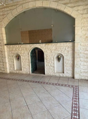 Um arco de pedra que dá acesso a um quarto com chão de azulejos e uma bordadura estampada.