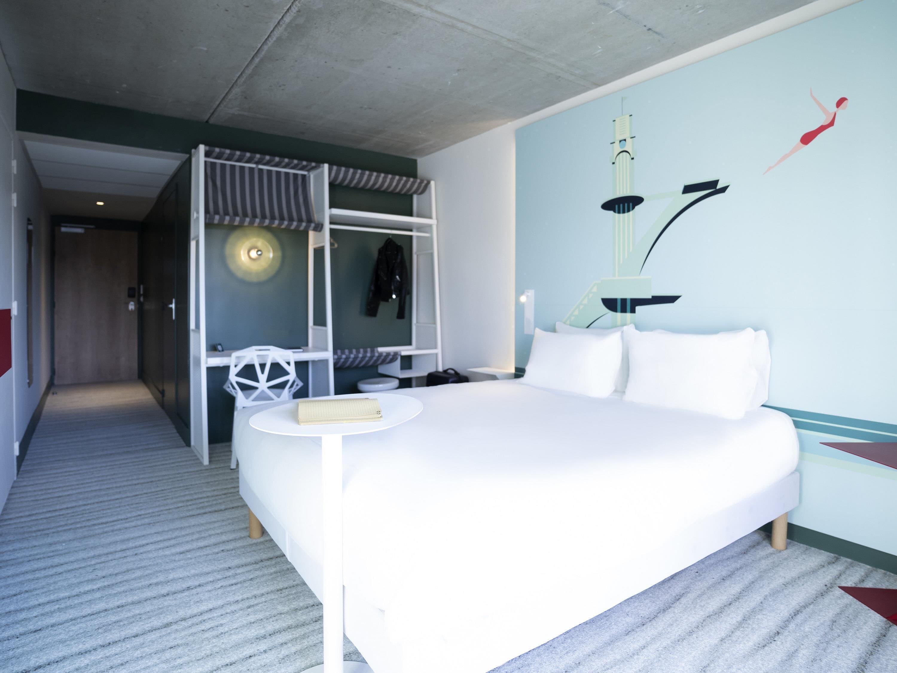 A hotel room with a large bed, a desk, and a chair. There is a coat rack and a lamp on the wall.