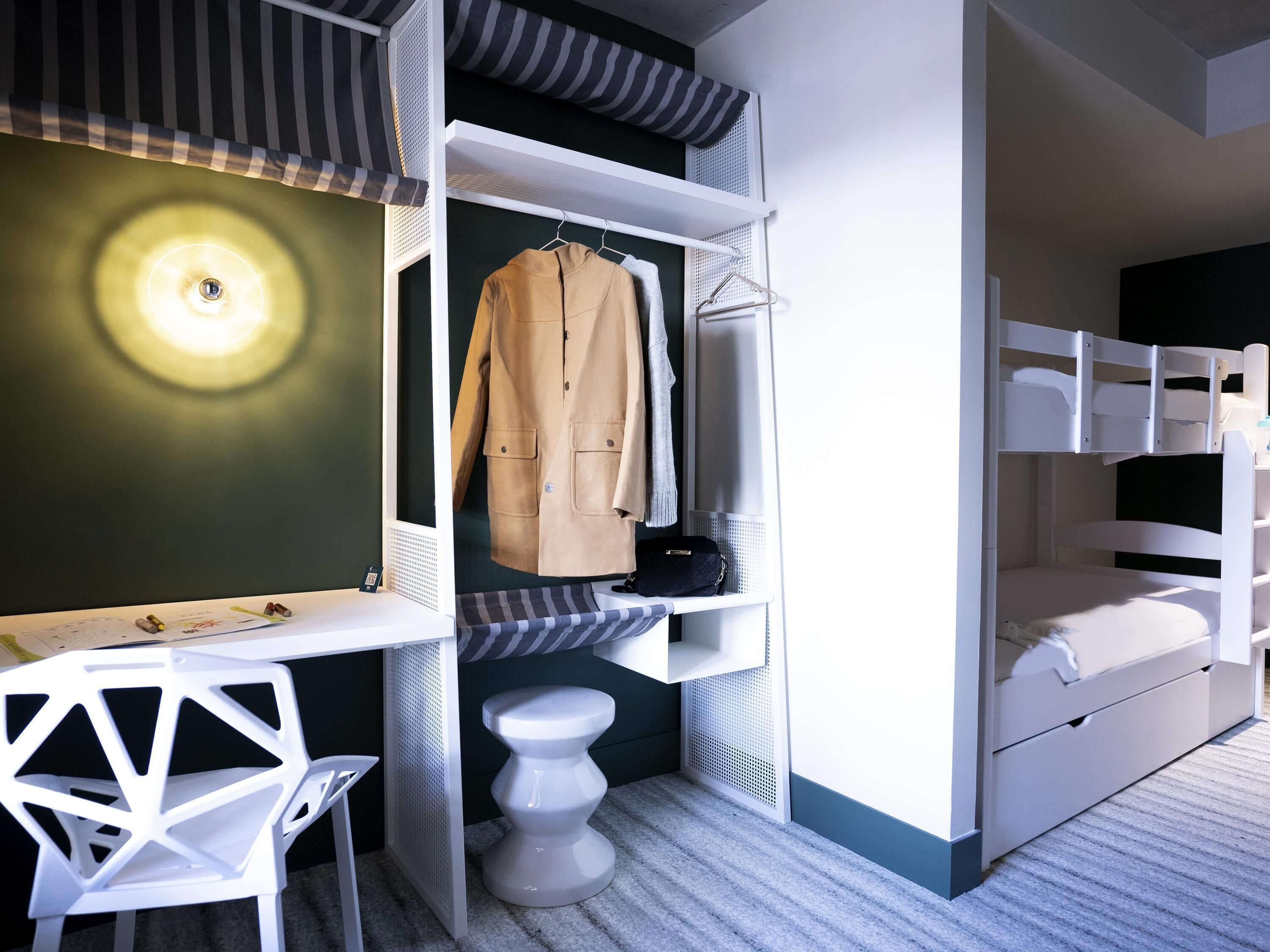 Une petite pièce comprenant un lit superposé, un bureau avec une chaise, un porte-manteau et un petit tabouret.