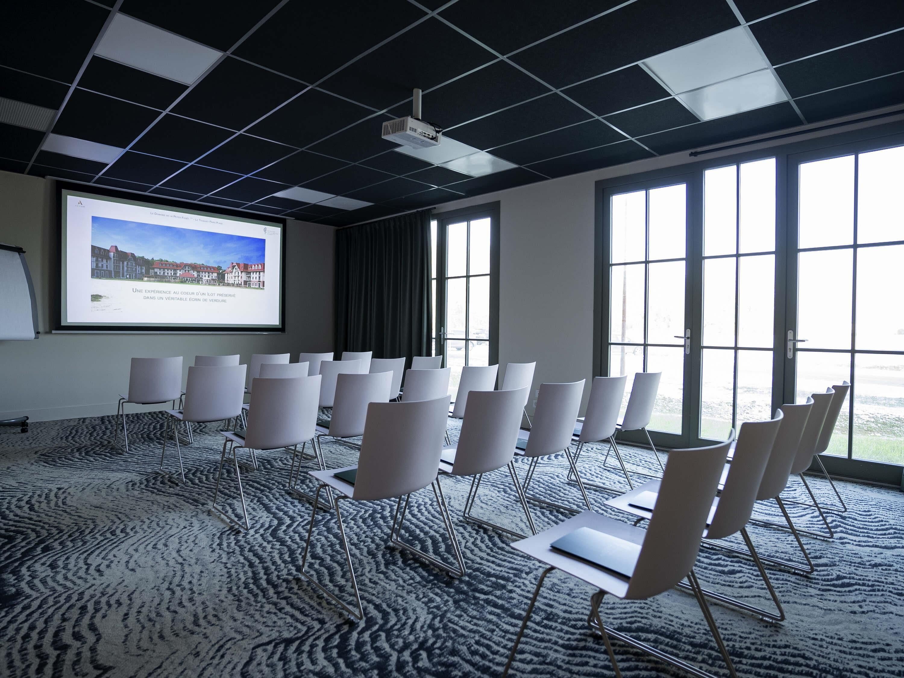 Een vergaderzaal met een projectiescherm, rijen stoelen, grote ramen en een kamerbreed tapijt met een patroon.