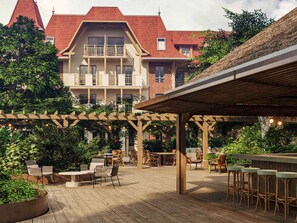 A wooden deck with outdoor seating, surrounded by lush greenery and a building with a red-tiled roof. - ibis Styles Le Touquet (Le Touquet-Paris-Plage)