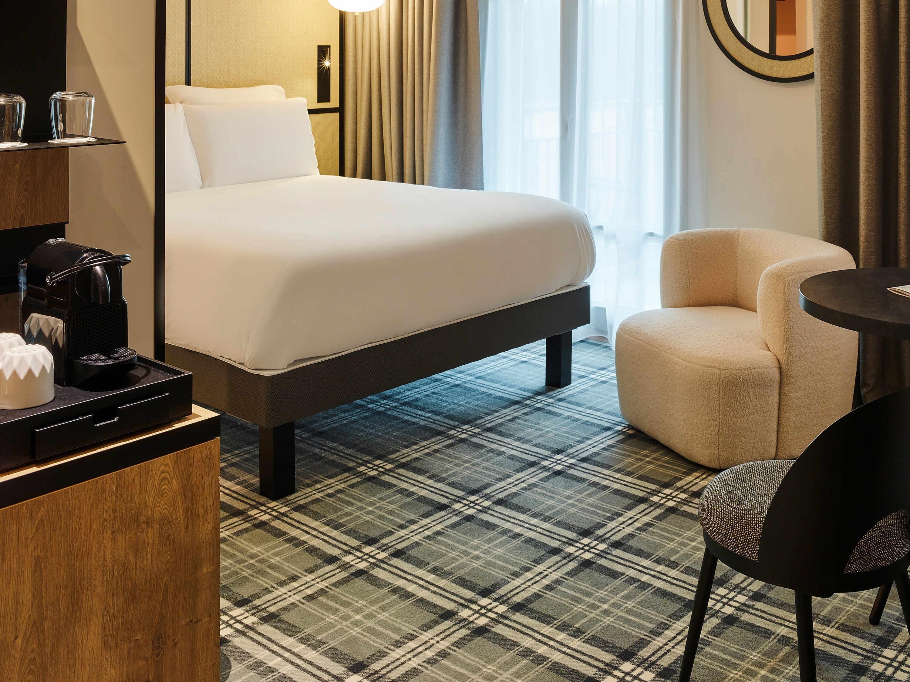 A hotel room with a bed, a chair, a small table, and a coffee maker on a wooden dresser.