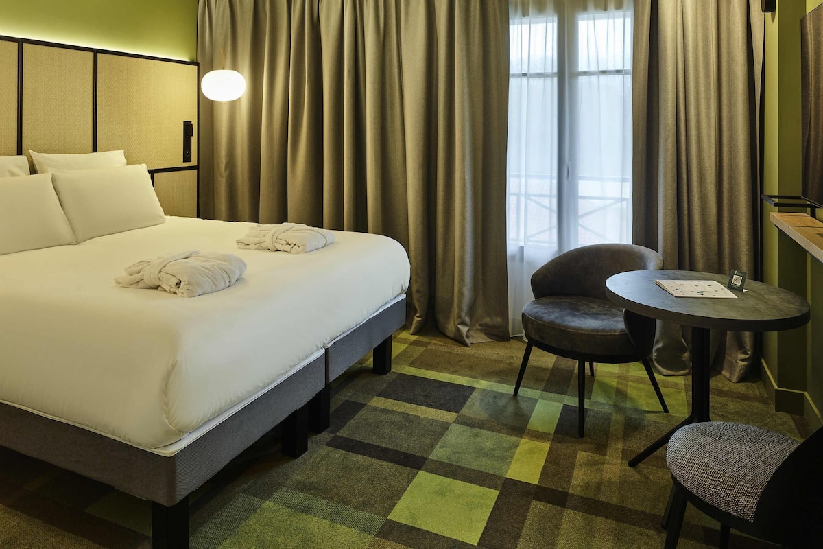 Hotellihuone, jossa on sänky, tuoli, pieni pöytä ja näkymä ulos.