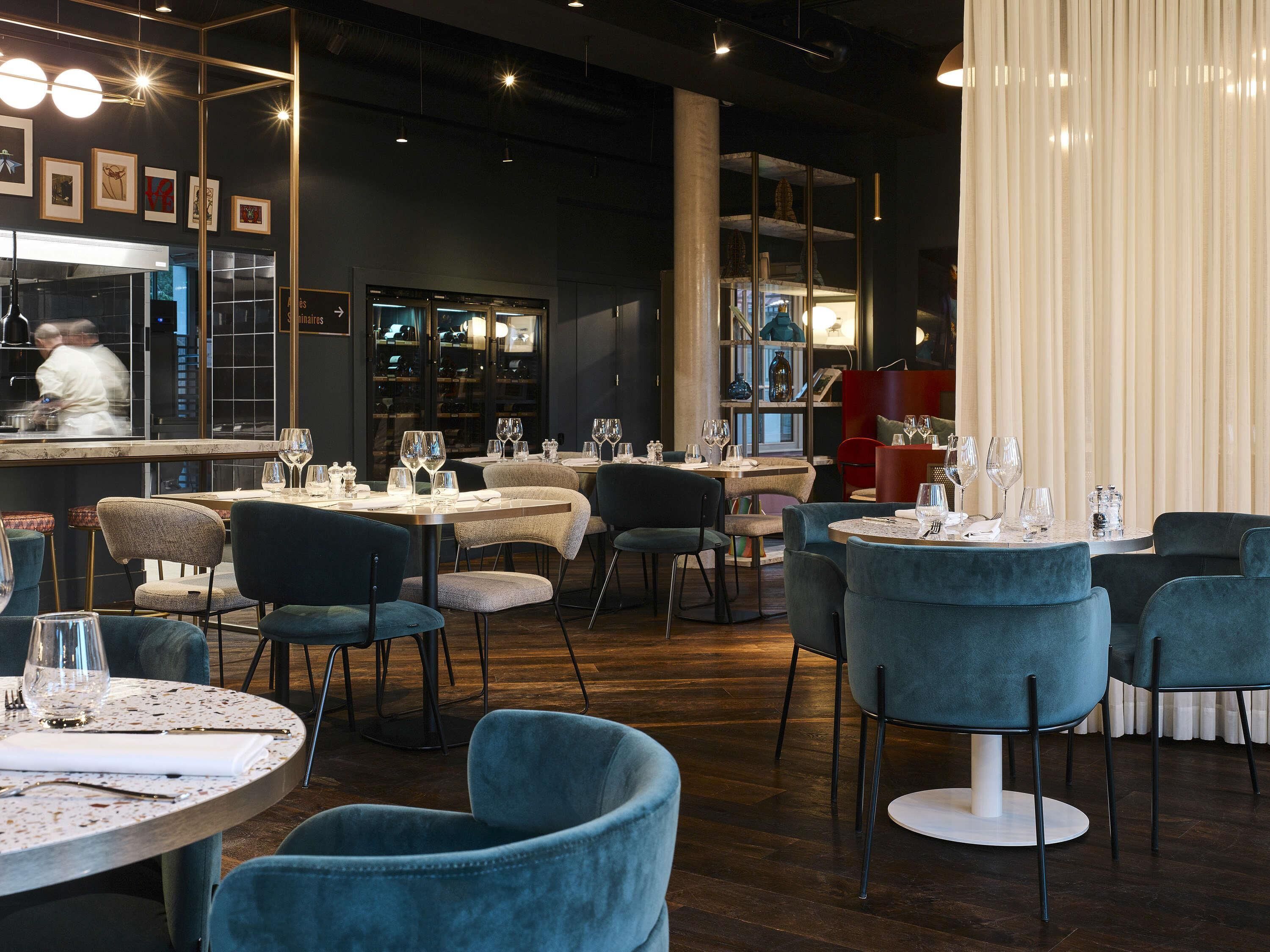 Un intérieur de restaurant moderne avec des tables rondes, des chaises recouvertes de tissu bleu et un bar où un chef prépare les plats.