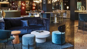 Un espacio lounge moderno con un sofá azul oscuro, otomanas redondas y una mesita de centro. Se observa una zona de bar al fondo con un taburete alto y un espejo.