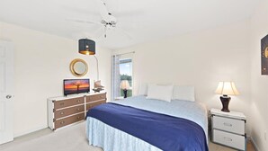 A bedroom with a bed, a dresser, a nightstand, a ceiling fan, a window with curtains, and a wall-mounted TV. - Serene Sunrise & Shimmering Skyline. 5 Star Hosts (Ocean City)