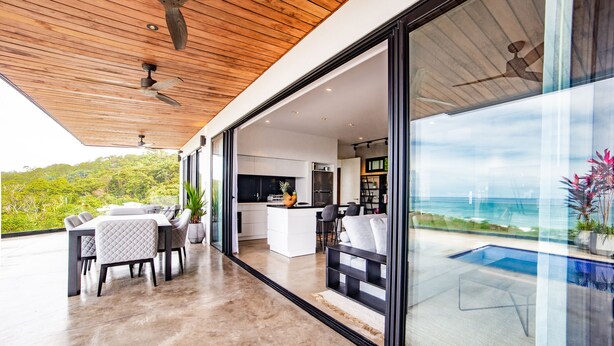 Un área de estar moderna y de concepto abierto con cocina, comedor y un amplio ventanal que da a un balcón con piscina y vista al océano.