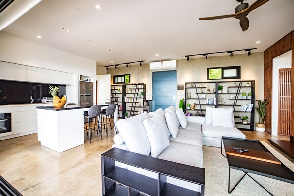 A modern living room with a sofa, coffee table, and a TV. There is a kitchen area with a fridge, stove, and dining table.