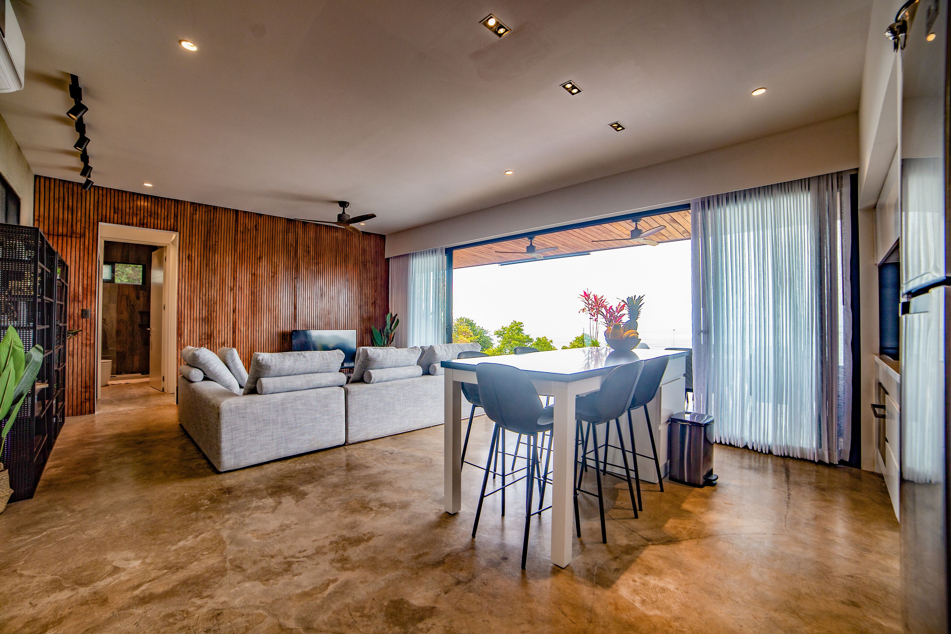 Una sala de estar moderna con un sofá, una mesa de comedor y sillas. Hay un ventilador de techo, una ventana con cortinas y vistas a zonas verdes.