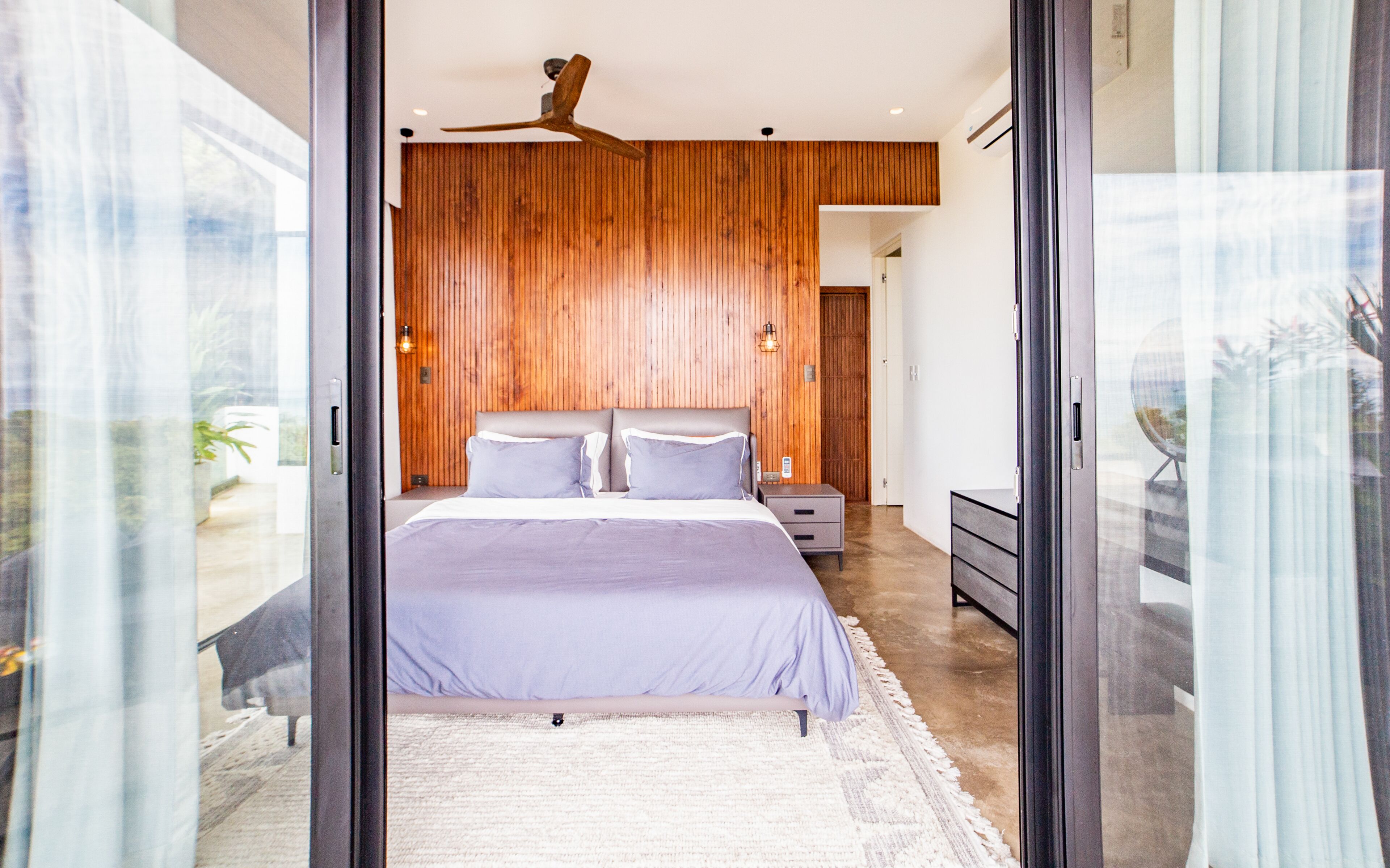 Un dormitorio moderno con pared de madera, una cama con colcha gris, una mesita de noche y un ventilador de techo.