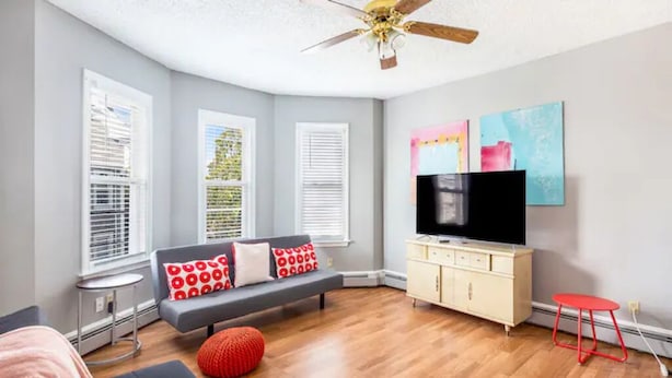 A modern living room with a grey sofa, red and white patterned cushions, a red ottoman, a television on a white cabinet, and a ceiling fan. - 5⭐️BEST PRICE⭐️ Whole apt.3 bdrm for 2-8, long term (Providence)