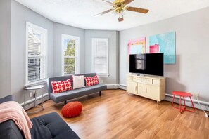 A modern living room with a grey sofa, red and white patterned cushions, a red ottoman, a television on a white cabinet, and a ceiling fan.