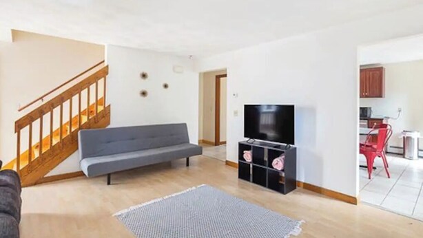 A living room with a grey sofa, a TV on a black stand, a wooden staircase, and a kitchen area with red chairs. - 5⭐️BEST PRICE⭐️Robert's 3 Room for 2-7ppl Long term (Providence)