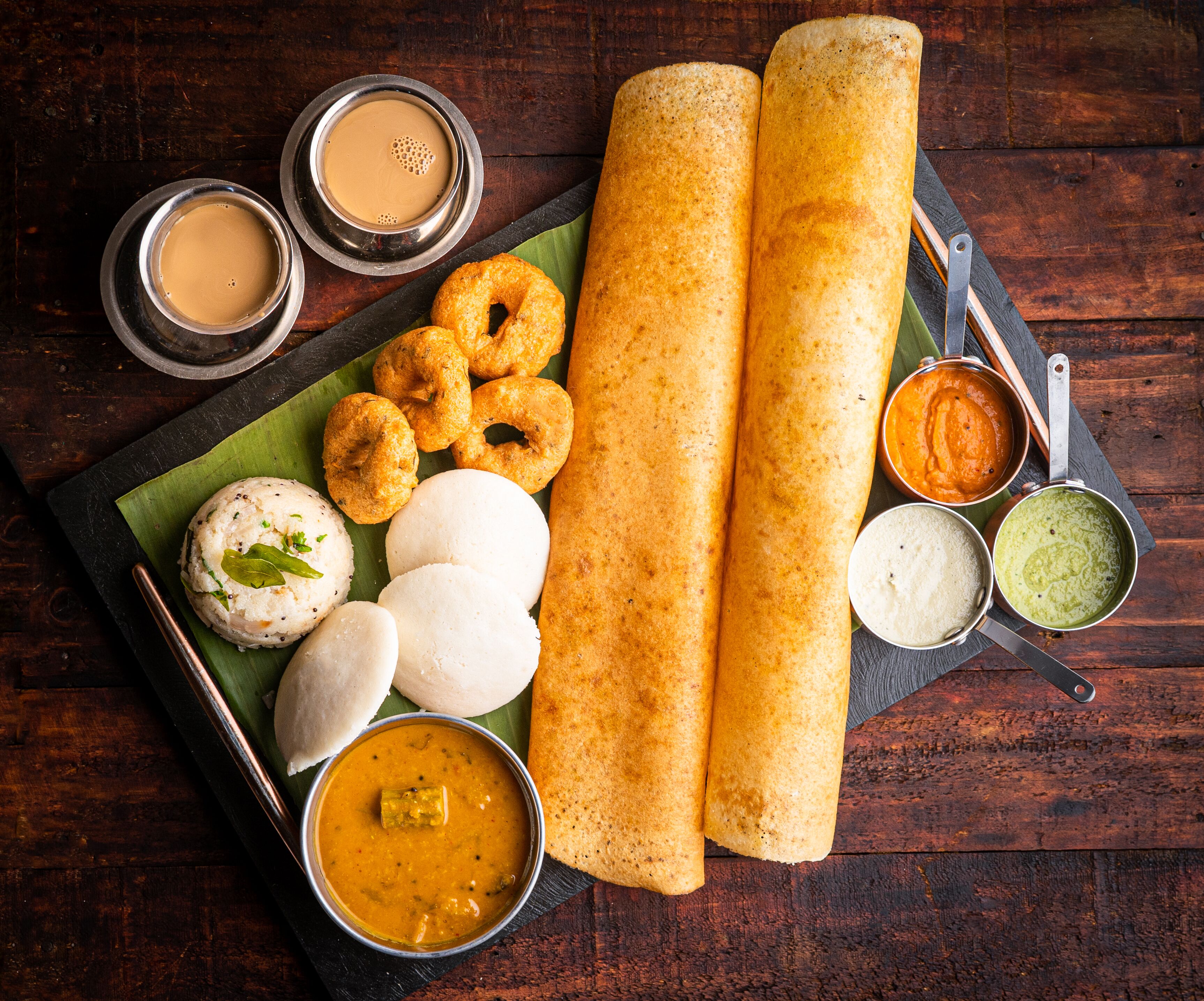 A variety of South Indian dishes including dosas, idlis, and chutneys on a wooden table.