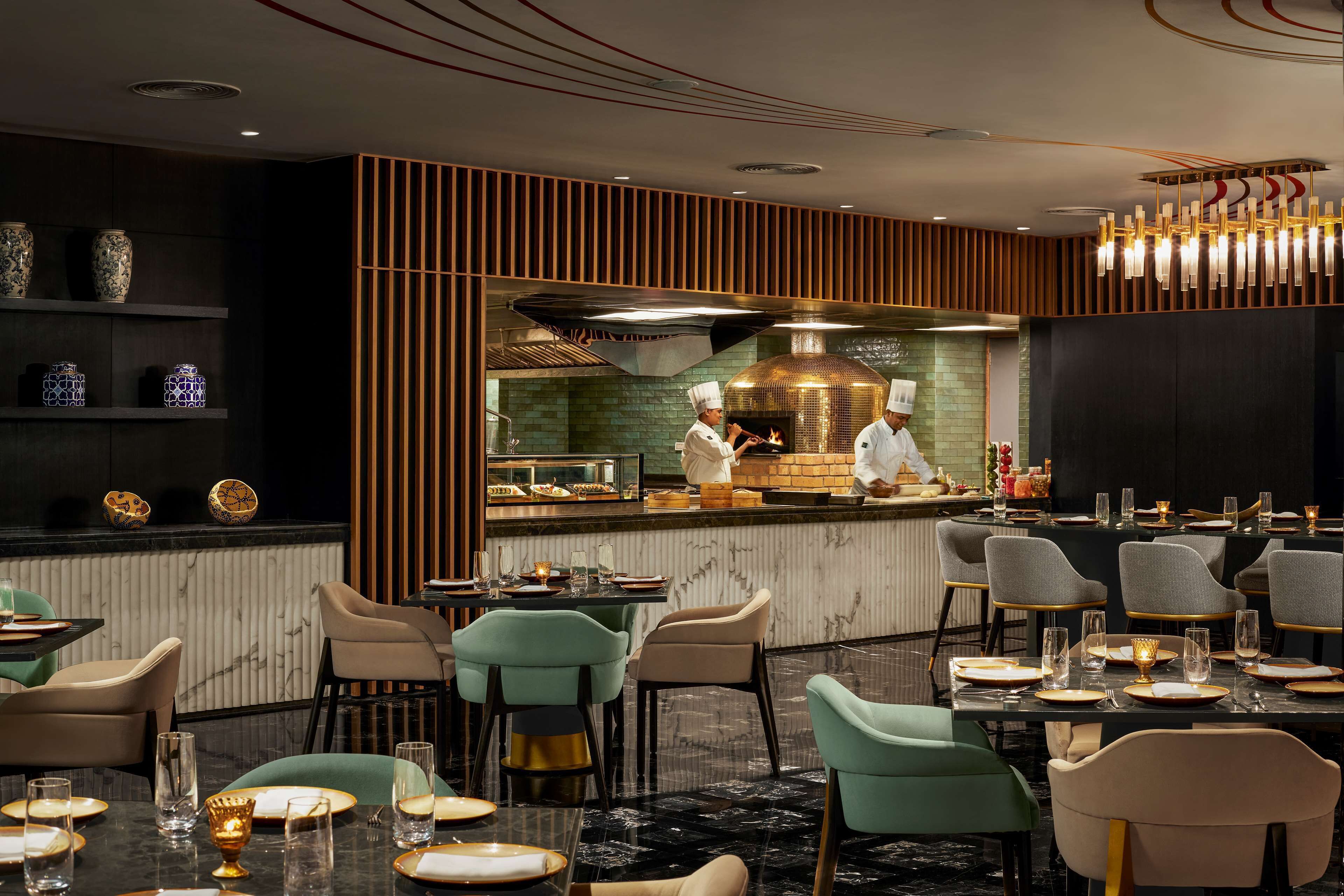 Un restaurante con una zona de cocina central, varias mesas con sillas y jarrones decorativos en estanterías.