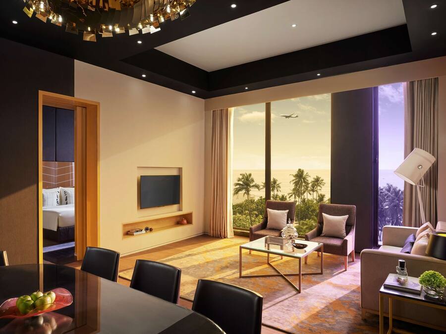 Habitación de hotel moderna con un ventanal grande, televisión de pantalla plana, zona de comedor y área de estar con vistas a palmeras.