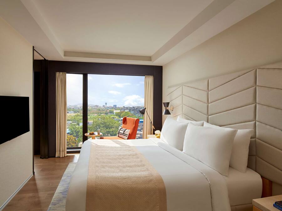 A hotel room with a large bed, a flat-screen TV mounted on the wall, a nightstand, and a view of the city through the window.