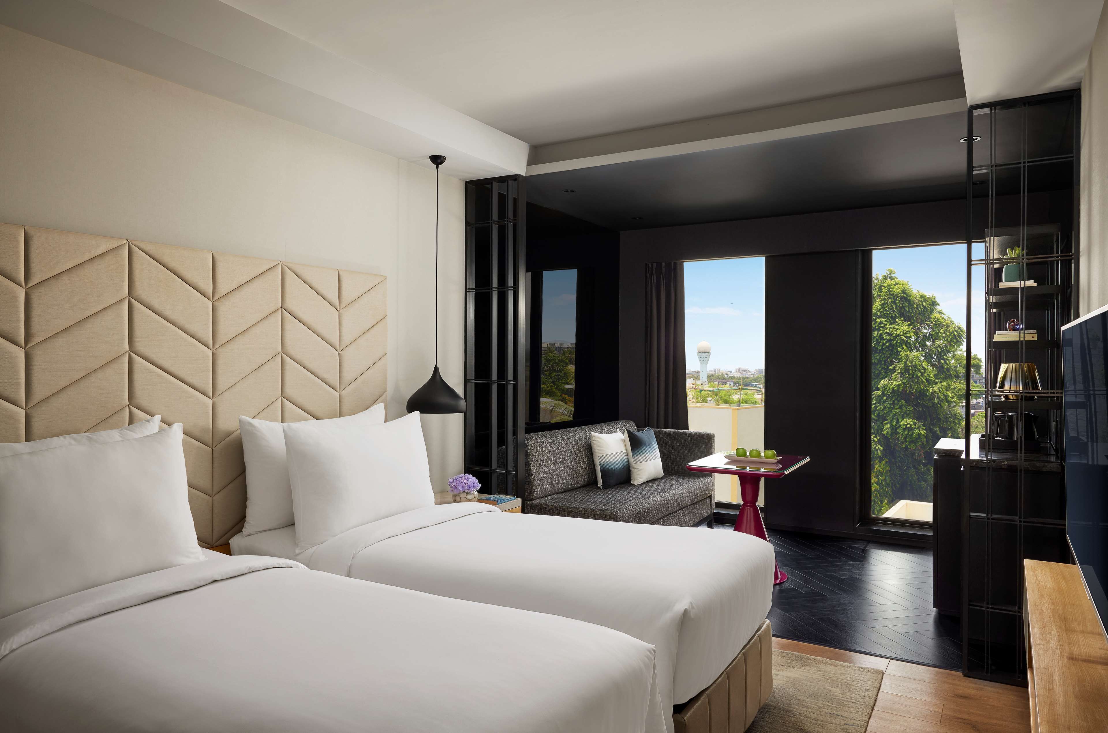 A modern hotel room with two beds, a sofa, a small table, and a large window offering a city view.