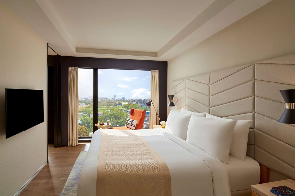 Ein Hotelzimmer mit einem großen Bett, einem Flachbildfernseher, einem Nachttisch, einer Lampe und Blick auf die Stadt.