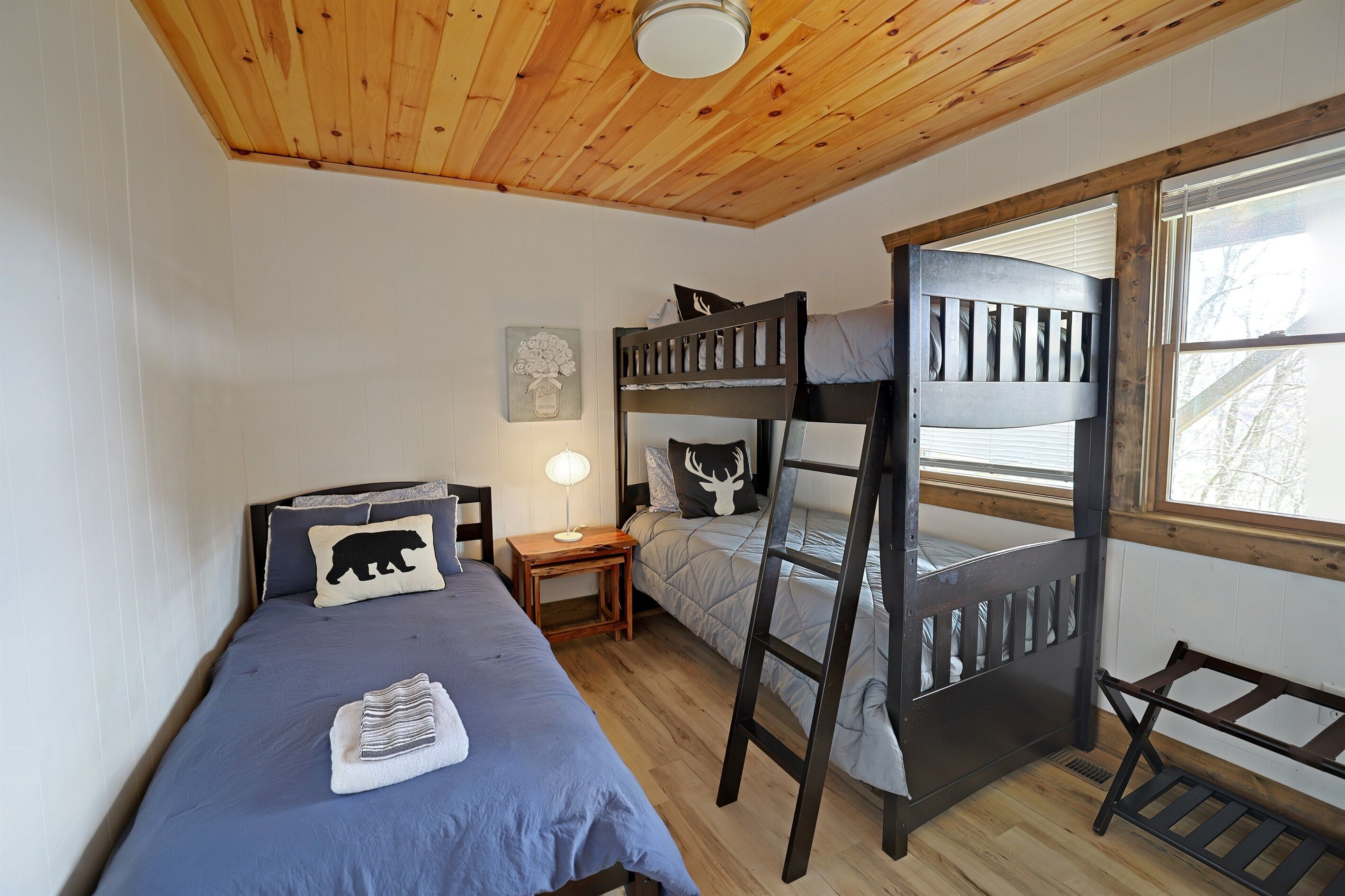 A bunk bed room with a wooden ceiling, a nightstand, a lamp, and a window with a view of trees.