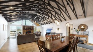 A spacious living area with a wooden dining table, comfortable seating, a stone fireplace, and large windows offering a view. - Ski Chalet: 0.5 mi to slopes. Great view-Fireplace-central A/C (Sugar Mountain)