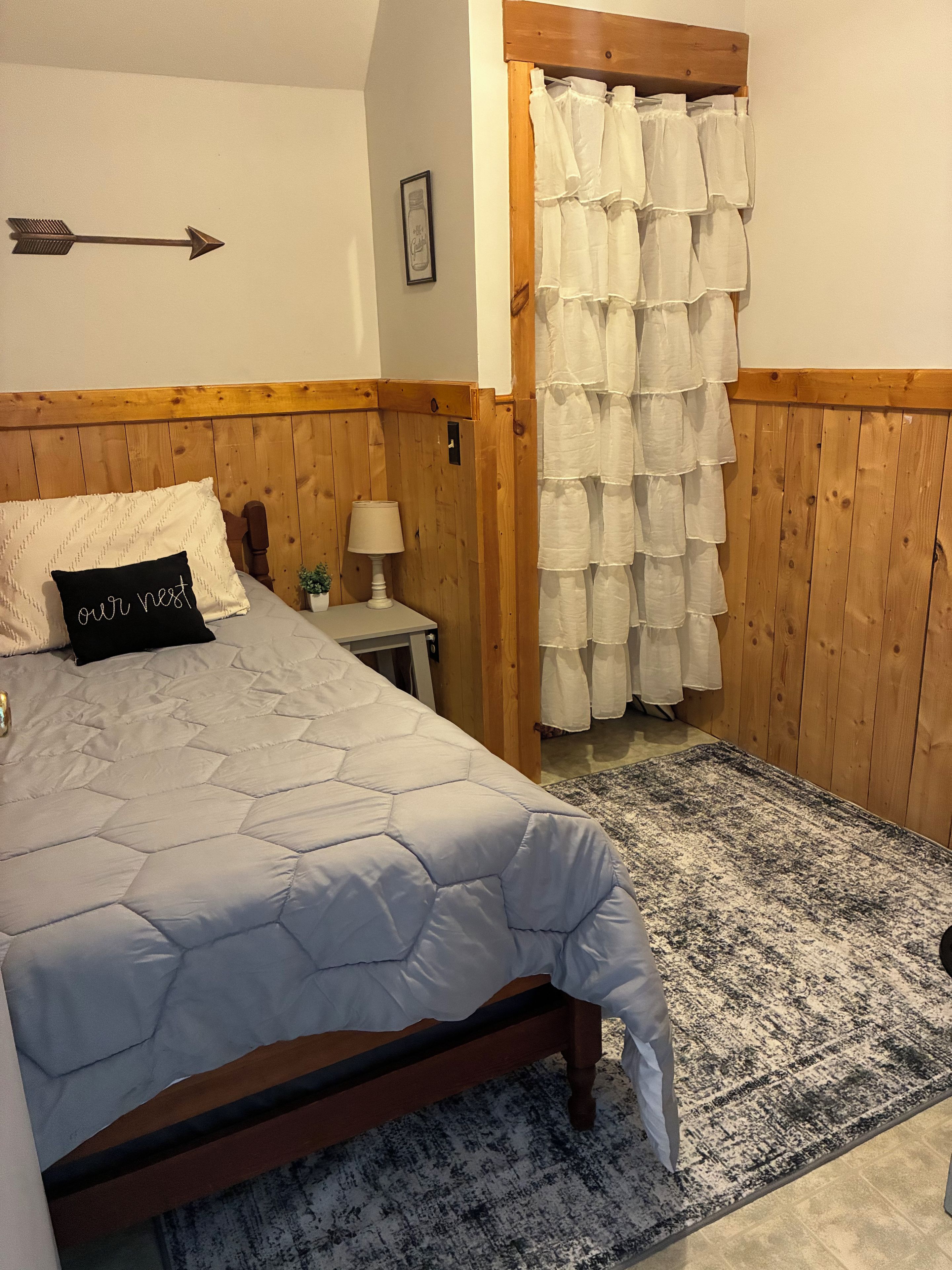 A bedroom with a bed, a nightstand, a lamp, a wooden headboard, a wall-mounted arrow decoration, and a white curtain.