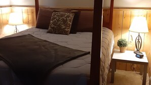 Ein Bett mit Baldachin, ausgestattet mit einer dunkelgrauen Tagesdecke, einem Holzkopfstück und einem Nachttisch mit Lampe und Wecker.
