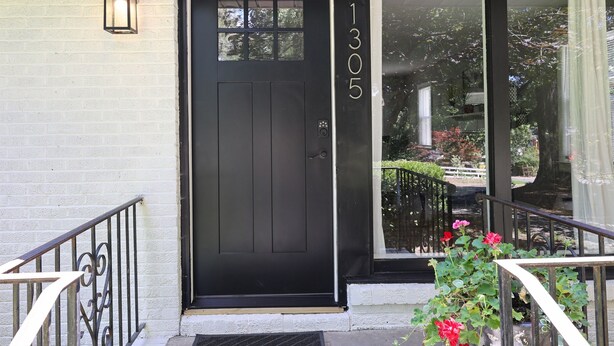 Une porte d’entrée noire portant le numéro 1305, une petite fenêtre au-dessus, et un pot de fleurs avec des fleurs rouges devant.