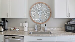 Une cuisine avec des meubles blancs, un évier en acier inoxydable, une machine à café, un four à micro-ondes et un miroir décoratif en forme d’arbre.